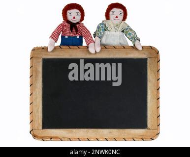 Raggedy Ann & Raggedy Andy tenere un cartello di lavagna per la vostra nota, messaggio, annuncio. Ritaglio isolato per voi. Le bambole di mini straccio si tengono sulla tavola. Foto Stock