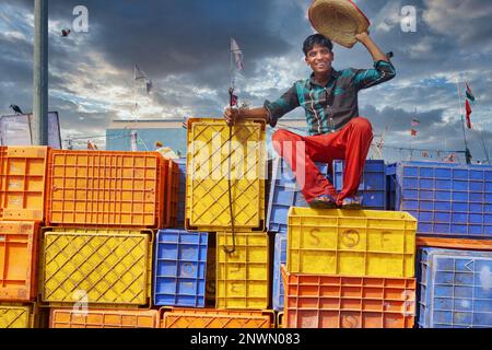 Un giovane indiano seduto su colorate casse di plastica nel Vecchio Porto di Mangalore, Karnataka, India del Sud, sollevando il cappello in un saluto allegro Foto Stock