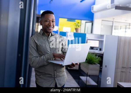 Ritratto di un uomo d'affari afro-americano che tiene in mano un computer portatile e sorride in ufficio. Lavorare in ufficio, business, finanza e concetto aziendale. Foto Stock