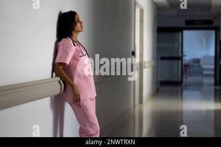 Medico di sesso femminile biraciale esausto con stetoscopio appoggiato alla parete in ospedale. Concetto di salute, reparto ospedaliero e assistenza. Foto Stock