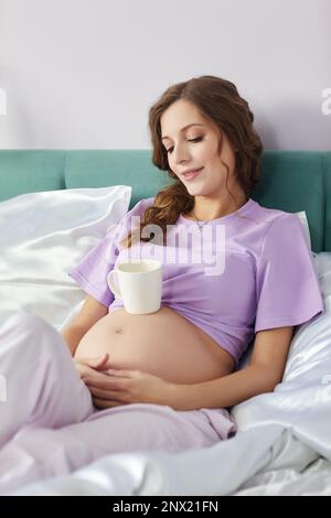 Una donna incinta felice si trova nel letto della sua camera da letto domestica e sorridente. Donna vestita di pajama godendo il suo stato di gravidanza. Una tazza di tè è sul Foto Stock