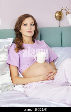 Una donna incinta si trova nel letto della sua camera da letto domestica. Donna vestita di pajama godendo il suo stato di gravidanza. Una tazza di tè è sul ventre. Foto Stock