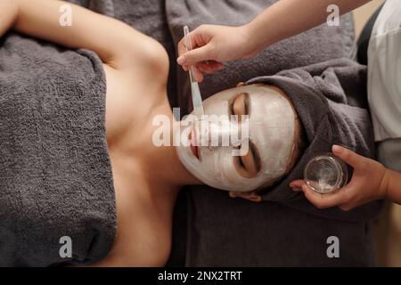 Cosmetologo applicare una maschera di argilla bianca brillante sul volto della giovane donna Foto Stock