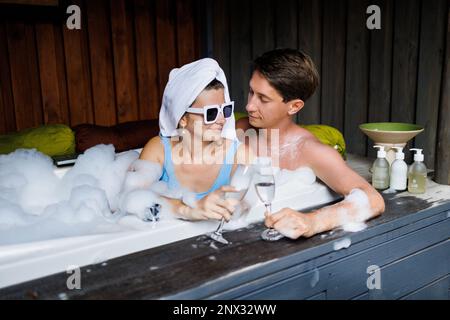 I giovani amanti del costume da bagno si crogiolano e baciano nella jacuzzi con schiuma nel centro benessere diurno. Il concetto di relazioni romantiche, passione, relax Foto Stock