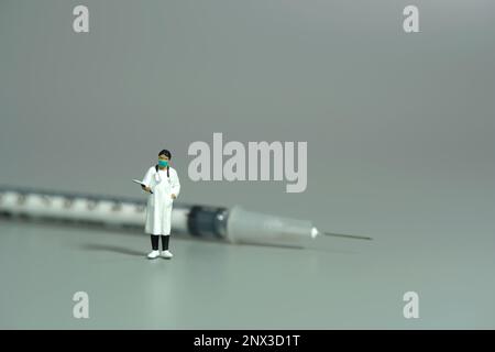 Fotografia di figura giocattolo delle persone in miniatura. Una dottoressa in piedi davanti alla siringa dell'ago su sfondo grigio. Foto dell'immagine Foto Stock