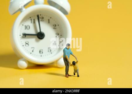 Fotografia di figura giocattolo delle persone in miniatura. Tempo per i bambini concetto. Un padre e un figlio in piedi davanti all'orologio bianco. Sfondo giallo isolato. IMA Foto Stock