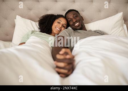 Giovane coppia multirazziale sorridente sdraiata a letto e tenuta le mani Foto Stock
