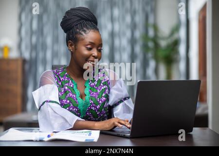 Studentessa universitaria africana che fa i compiti, studia i corsi in linea, scrivendo il progetto di tesi con le note a casa, seduta al tavolo. Foto Stock