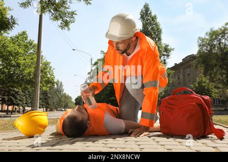 Lavoratore con una bottiglia d'acqua che aiuta un collega sulla strada cittadina. Che soffre di colpo di calore Foto Stock