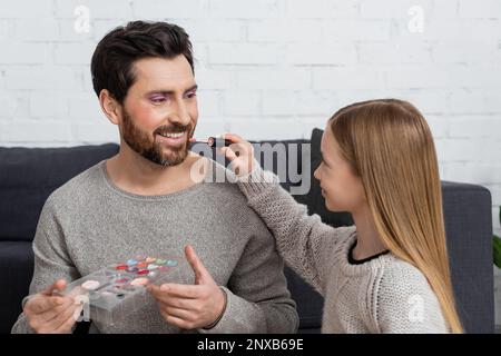 ragazza felice che applica la lucentezza del labbro sulle labbra del padre che tiene la tavolozza di trucco, immagine di riserva Foto Stock