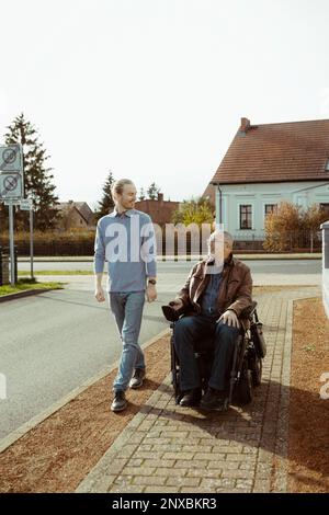 Uomo anziano sorridente con disabilità in sedia a rotelle motorizzata da parte di un giovane caregiver che cammina sulla strada durante le giornate di sole Foto Stock