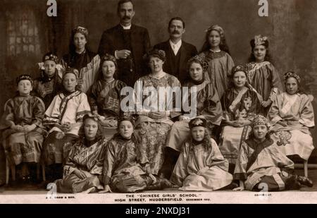 L’Estremo Oriente arriva nel West Yorkshire, Inghilterra, Regno Unito: Le alunne della ‘Chinese Girls’ School’ di Huddersfield High Street si siedono nel dicembre 1912 per un ritratto di gruppo con i loro insegnanti e un chierico vestito esoticamente in costume cinese tipico dell’epoca. Cartolina d'epoca dalle tonalità seppie pubblicata da John Edward Shaw & Son, fotografi di Huddersfield e Sheffield. Foto Stock