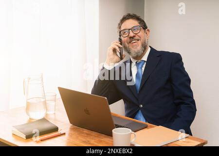 Un uomo d'affari di mezza età con barba bianca e occhiali è seduto alla sua scrivania con un computer portatile. Sorride e parla al telefono, vestito da un su Foto Stock