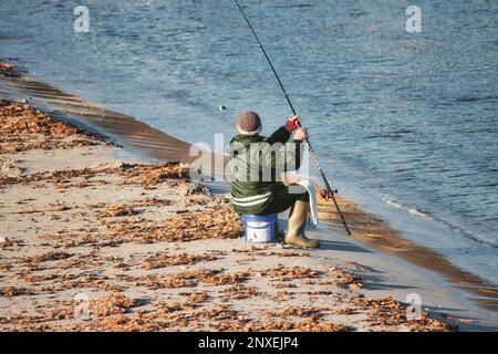 Un uomo anziano in una spiaggia di sabbia si sedette su un secchio di pesca a mosca con una canna da pesca sulla costa Foto Stock