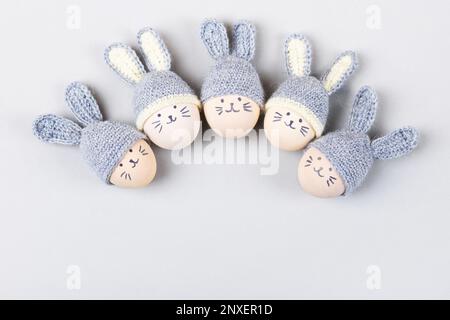 Cinque uova di Pasqua con volti divertenti e cappelli grigi uncinati con orecchie conigliate su sfondo grigio. Buon concetto di Pasqua. Biglietto d'auguri. Foto Stock