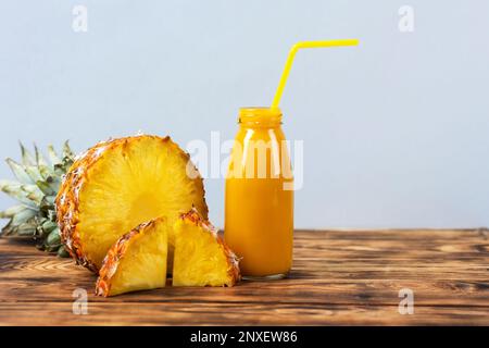 Fette di succosa ananas e un frullato fresco e sano in una bottiglia di vetro e una paglia su un tagliere di legno su uno sfondo di legno. Sano, disintossicante Foto Stock
