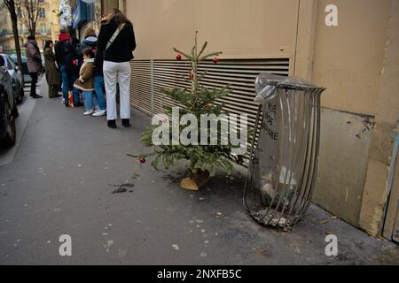 Albero di Natale lasciato accanto a una spazzatura può vicino a un ristorante popolare con una coda in strada Foto Stock