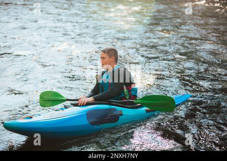 Atleta sportivo maschile che si addica con attenzione sulle rischiose, schiumose rapide e spruzzi nel suo kayak blu Foto Stock