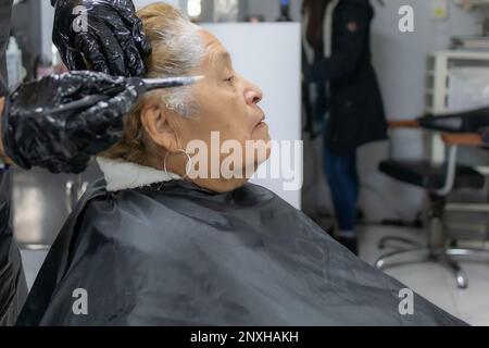 Stilista che inizia ad applicare colorante ai capelli corti di una donna anziana contro sfondo sfocato, espressione seria, vista laterale. Moda antica e bellezza c