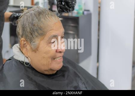 Stilista che applica il colorante ai capelli corti di una donna anziana con sfondo sfocato, espressione con un sorriso leggero, vista laterale. Moda e bellezza