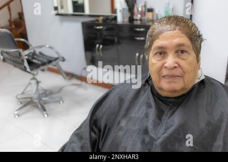 Vecchia donna con tintura sulla testa seduta su una sedia in salone di bellezza con uno sfondo sfocato, capelli corti, espressione con un sorriso leggero, vista frontale. O