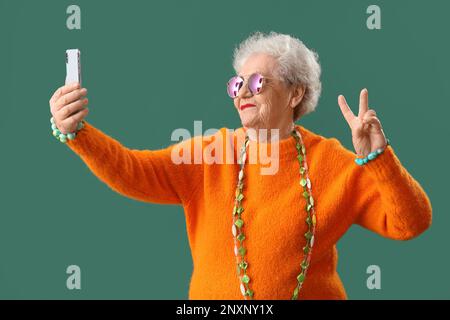 Donna anziana con accessori luminosi che prendono selfie su sfondo verde Foto Stock