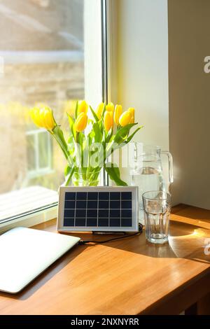 Portatile pannello solare di carica computer portatile, caraffa d'acqua e vaso con tulipani sul tavolo vicino alla finestra Foto Stock