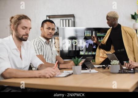 Giovane uomo africano in camicia a strisce sorridente mentre guardava la macchina fotografica, seduto con un collega caucasico maschio alla scrivania. Gli economisti passano il tempo in carica per lavorare al rapporto finanziario. Foto Stock