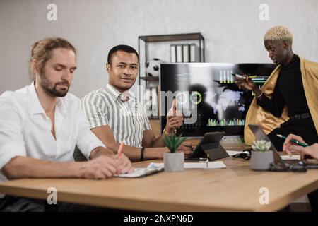 Giovane uomo africano in camicia a strisce sorridente mentre guardava la macchina fotografica, seduto con un collega caucasico maschio alla scrivania. Gli economisti passano il tempo in carica per lavorare su un rapporto finanziario che mostra il pollice in su Foto Stock