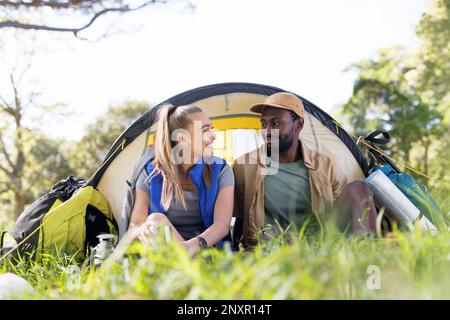 Felice, coppia diversa campeggio nella foresta seduto di fronte alla tenda, sorridendo l'uno contro l'altro Foto Stock