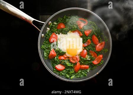 Spinaci, pomodori e uova fritte in una padella fumante su una stuuppa nera, cucinare un pasto sano per colazione o pranzo, vista dall'alto, copia Foto Stock