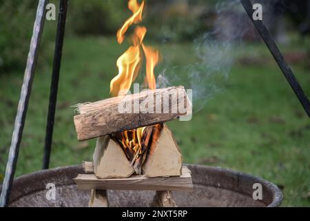 Legna da ardere accatastata intelligente tronchi che bruciano con fiamme e fumo in una ciotola di fuoco sotto un treppiede grill in un giardino per una festa barbecue, spazio copia, selezionato Foto Stock