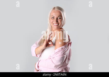 Donna matura felice mostrando braccio con bendaggio dopo la vaccinazione e gesto OK su sfondo chiaro Foto Stock