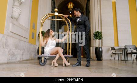 Coppia elegante all'ingresso dell'hotel. Azione. Donna elegante siede sul carrello europeo dei bagagli negli hotel. Coppia di lusso bellissima in un hotel costoso. Foto Stock