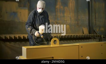 L'operaio in tuta dipinge la parte metallica con la pistola a spruzzo. Fermo. Verniciatura a spruzzo di parti metalliche di grandi dimensioni in fabbrica. Verniciatura di grandi oggetti metallici o parti in fabbrica Foto Stock