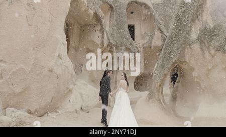 Atmosfera magica per la sposa e lo sposo in Cappadocia. Azione. Formazioni rocciose, neve caduta e bella donna in abito bianco e un uomo in sui nero Foto Stock