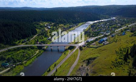 Veduta aerea di una valle pittoresca con un villaggio. Fermo. Ampio fiume attraversato da un ponte e colline boscose Foto Stock