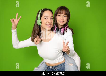 Divertente sorridente giovane donna e bambina ragazza sorelle indossando cuffie abbracciando guardando la fotocamera isolato su sfondo verde ritratto studio. Foto Stock