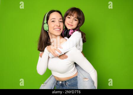 Divertente sorridente giovane donna e bambina ragazza sorelle indossando cuffie abbracciando guardando la fotocamera isolato su sfondo verde ritratto studio. Foto Stock