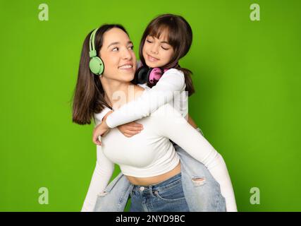 Divertente sorridente giovane donna e bambina ragazza sorelle indossando cuffie abbracciando guardare l'un l'altro isolato sul colore verde sfondo studio ritratto Foto Stock