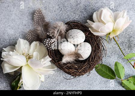 Vista dall'alto del concetto di biglietto di pasqua con nido pieno di uova e fiori di rosa bianchi su sfondo di cemento Foto Stock
