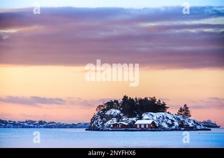 Vista panoramica di piccole case situate su ruvida scogliera isola vicino al mare calmo coperto di ghiaccio contro cielo nuvoloso tramonto in inverno a Grimstad in Agder co Foto Stock