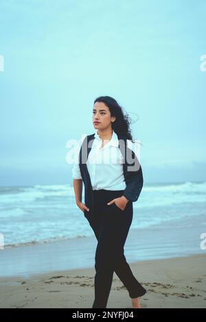 Giovane donna penosa in abiti bianchi e neri in piedi sulla spiaggia sabbiosa con le mani in tasca mentre si guarda lontano dal mare ondeggiante Foto Stock