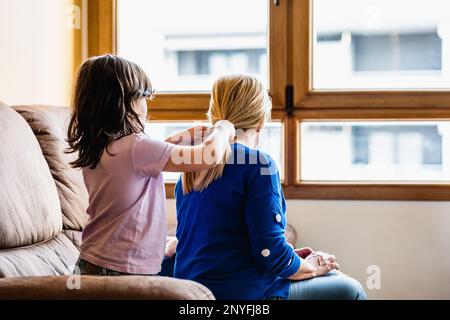 Vista laterale di una bambina concentrata seduta sul divano in pelle e spazzolando i capelli della madre contro la finestra nel soggiorno Foto Stock