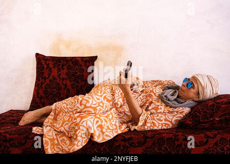 Corpo completo di donna musulmana anziana in abito arancione e turbante sdraiato sul divano con coperta rossa e cuscini mentre navigano sul telefono cellulare a casa in Mauritan Foto Stock