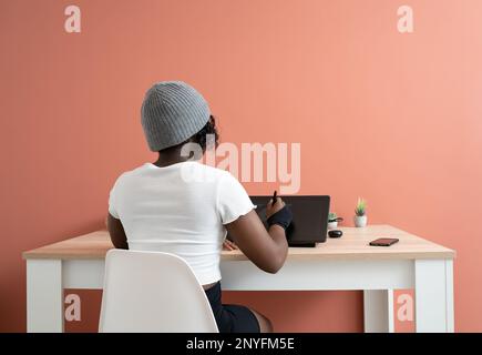 Vista posteriore di una giovane designer afro-americana in t-shirt bianca e cappello seduti al tavolo con stilo mentre disegnano su un tablet grafico Foto Stock