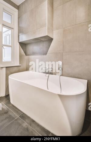 Vasca da bagno in ceramica bianca di forma ovale con rubinetto cromato collocato vicino alle pareti piastrellate beige in un bagno contemporaneo Foto Stock