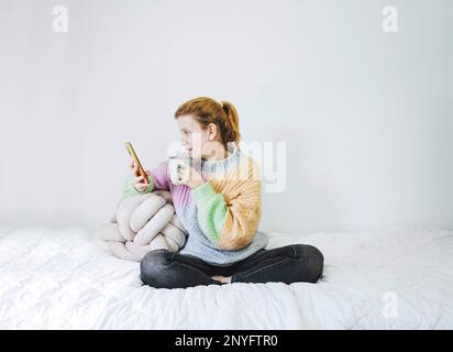 Corpo pieno di contenuti giovane donna in colorato maglione lavorato a maglia e jeans seduti su comodo letto in posizione lotus, mentre si gode una tazza di tè caldo e mes Foto Stock