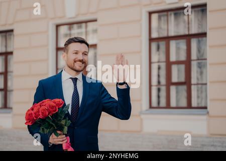 Bel giovane uomo positivo vestito in classico vestito blu e cravatta con camicia bianca saluto il suo partner di affari all'aperto, tenendo bouquet di rose rosse Foto Stock