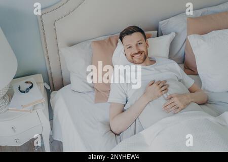 Giovane uomo ben riposato in maglietta bianca svegliarsi al mattino con sorriso, felice dopo aver dormito abbastanza, sdraiato in comodo letto mentre coperto di bl Foto Stock